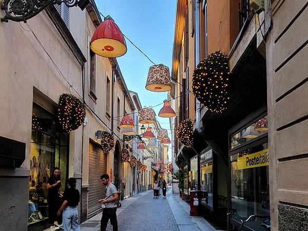 Via del centro storico di Parma con lampade decorative sospese e negozi caratteristici.