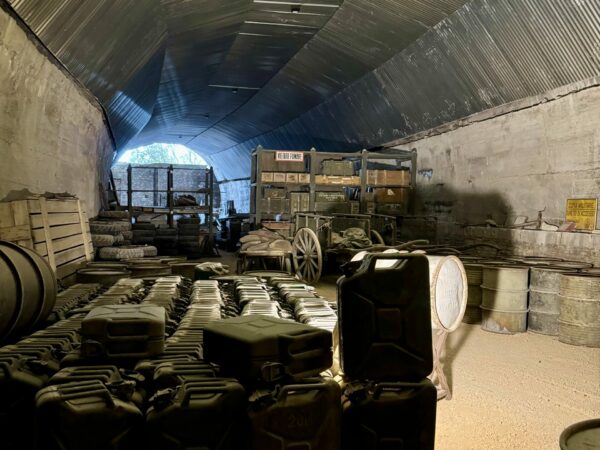 Galleria con soffitto in lamiera grecata contenente centinaia di taniche (jerrycans), barili e casse di munizioni.