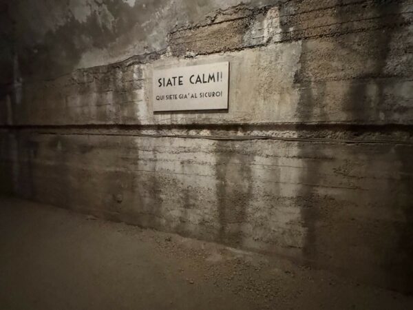 Dettaglio di una targa bianca su parete in cemento con la scritta nera "STATE CALMI! QUI SIETE AL SICURO".