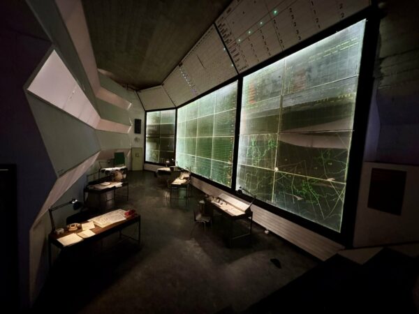 Sala operativa con enormi pannelli luminosi raffiguranti mappe geografiche e tabelle tattiche retroilluminate.