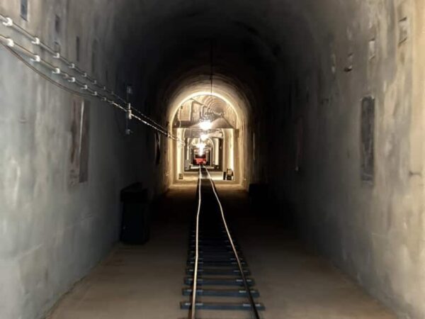 Prospettiva centrale dei binari a scartamento ridotto Decauville che si addentrano nel tunnel illuminato del bunker.